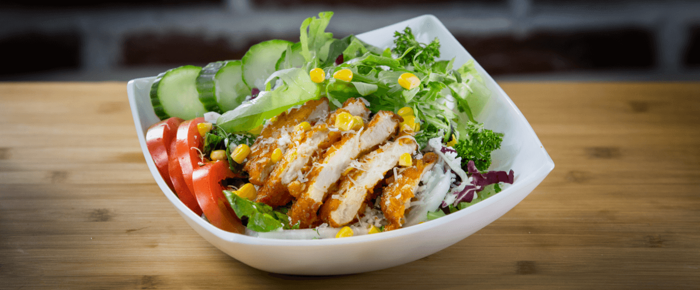 a white bowl filed with chicken salad consisting of grilled chicken, lettuce, sliced tomatoes, cucumber topped with corn & other garnishes