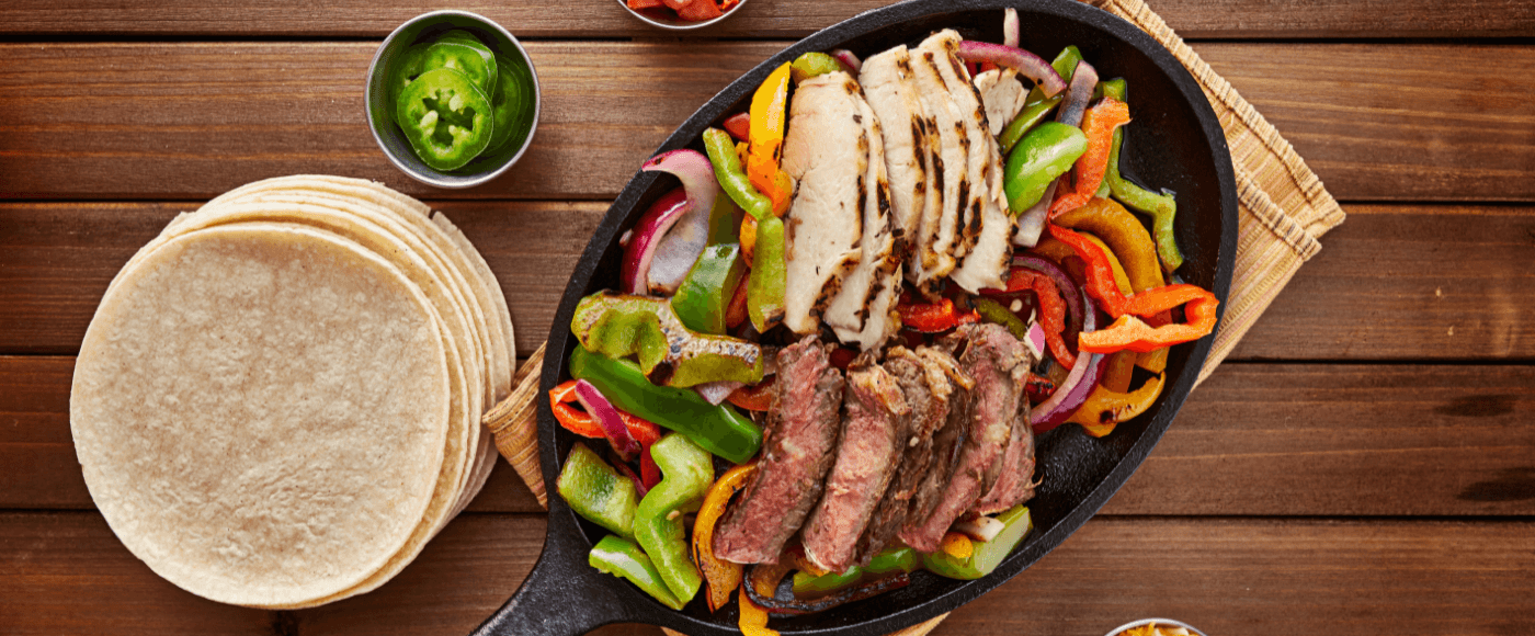a pan filled with chicken& steak fajitas placed on a towel on a rustic wooden table surrounded by tortillas, cheese & capsicum