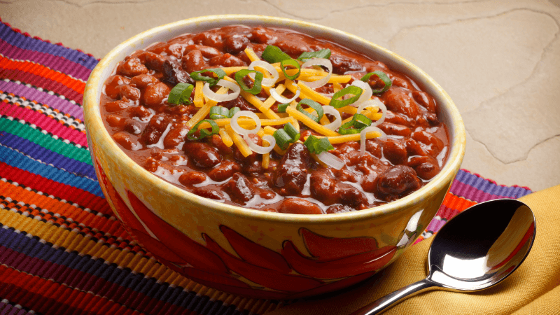 a colourful bowl of mexican slow-cooked bean chilli, garnished with cheese & green onions accompanied by a spoon