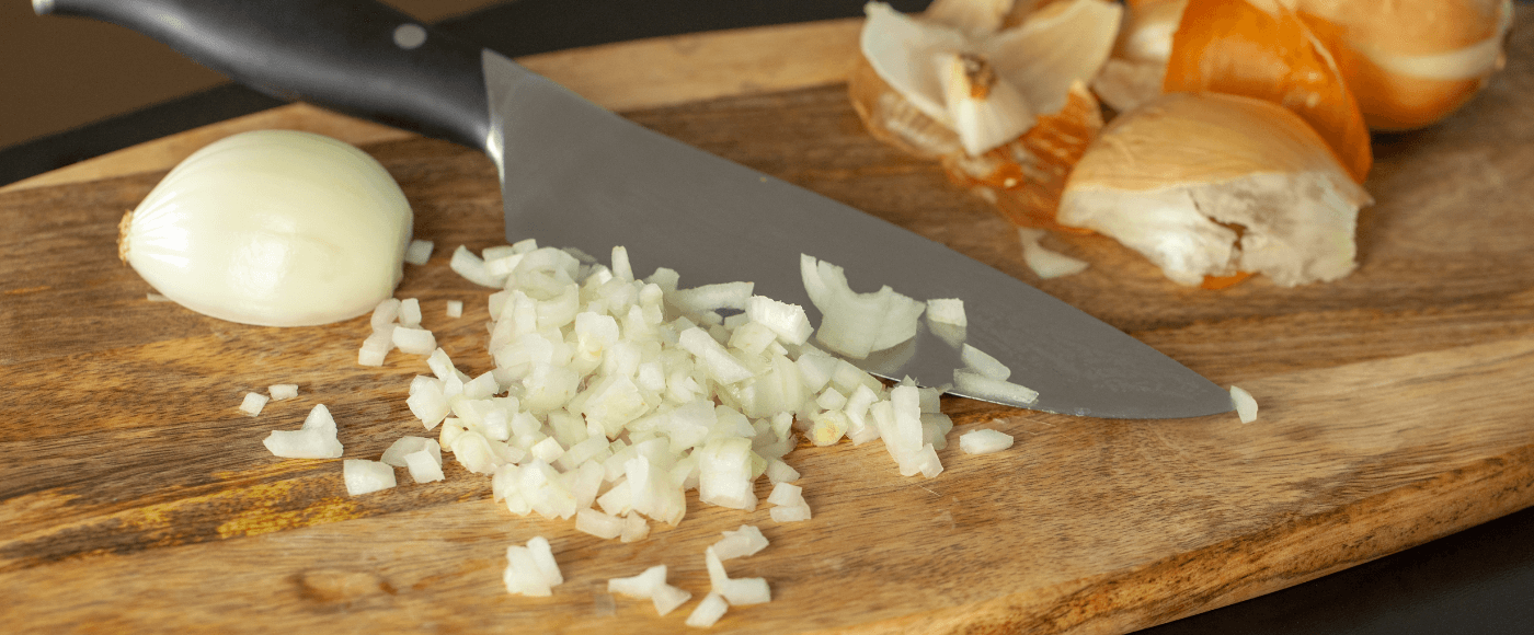 chopped onions on a wooden cutting board with kitchen knife, partially peeled onion & onions skins in the background
