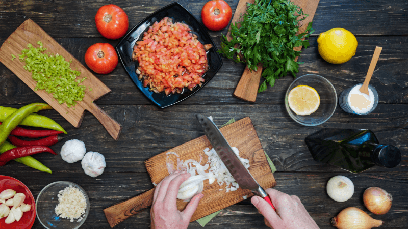 hands cutting onions with knife on cutting board, surrounded by assorted cut salsa ingredients on a rustic background