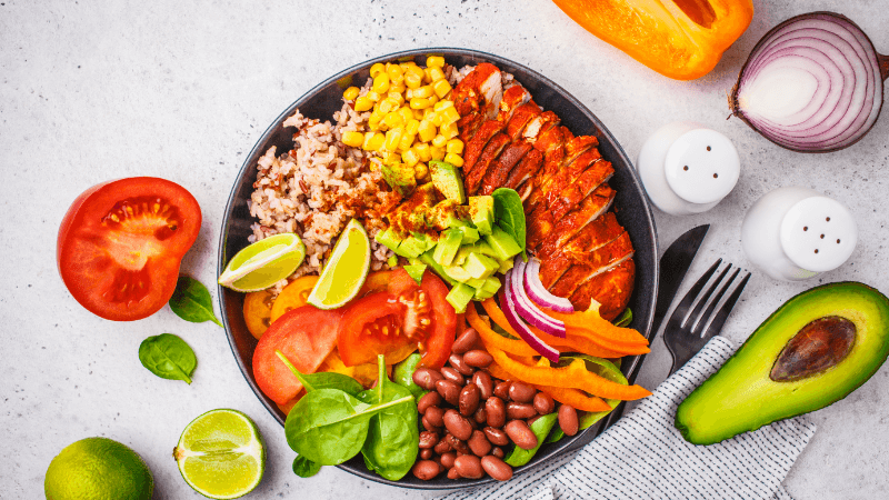 a vibrant bowl of ingredients such as chicken, beans, avocado, corn & fresh veggies styled with lime & seasonings