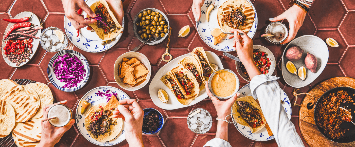 a table filled with tacos, toppings, sauces & hands assembling & sharing the meal