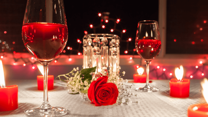 romantic dinner settings with two glasses of wine, a red rose & candles on a table, warm fairy lights in the background