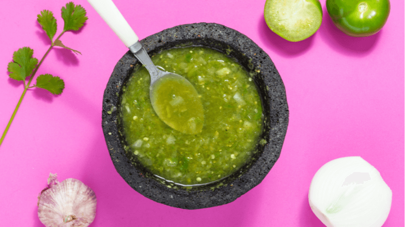 green salsa served in a black stone bowl with spoon surrounded by garlic, coriander, halved onion & a green tomato