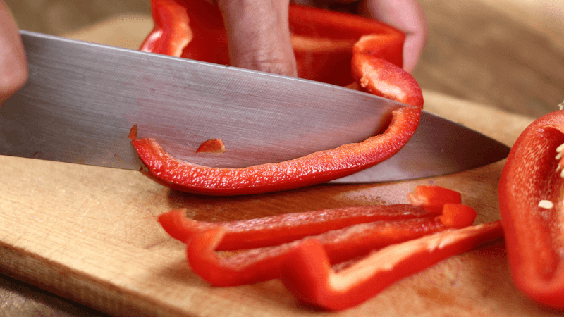 a hand slicing a red bell pepper with knife on a wooden cutting board
