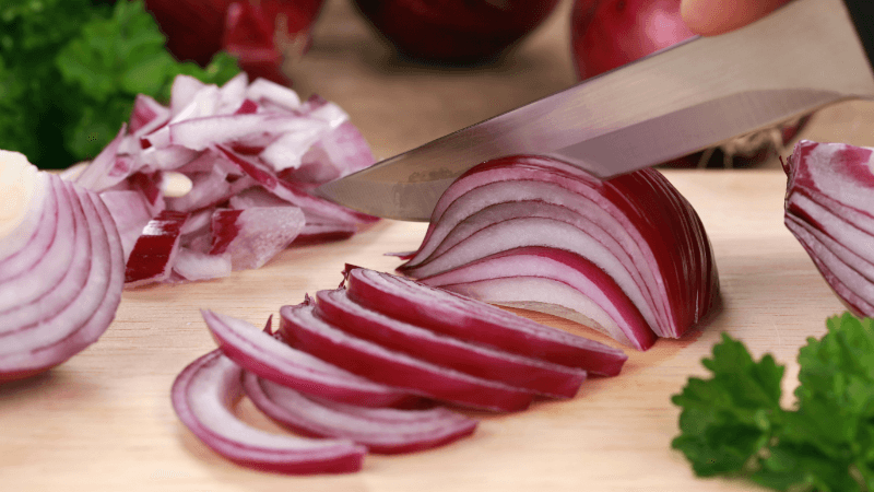 a hand slicing a red onion with knife on a wooden cutting board