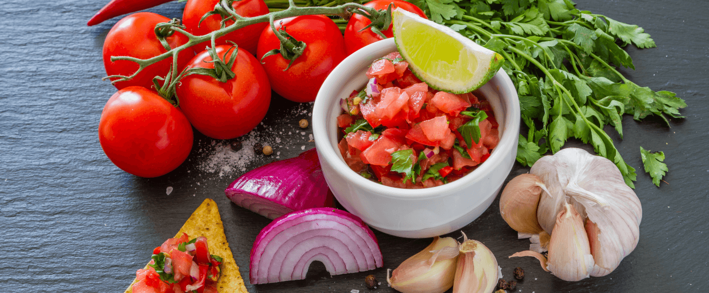 fresh salsa in a bowl with diced tomatoes, onions, cilantro, lime & spices, surrounded by fresh ingredients on a slate board