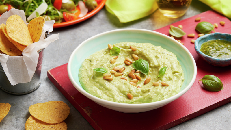 Smooth Pesto Guacamole topped with garnishes served i a light green bowl on a red wooden board