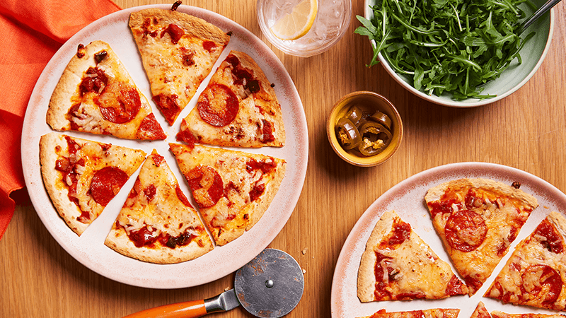 Two Easy Speedy Tortilla Pizzas with jalapeño on plates surrounded with pizza cutter & cilantro served on wooden table
