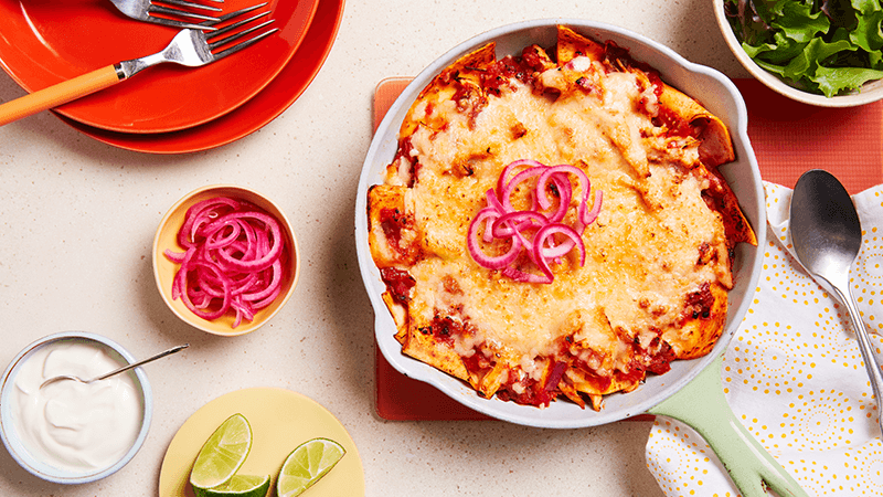 One Pan Chicken Enchiladas in a skillet surrounded with a bowl of greens, red onion, soured cream and lime wedges