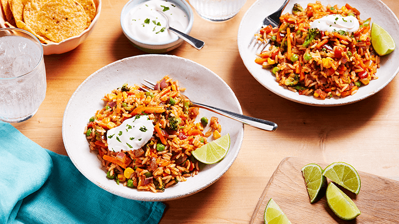 Two bowls of Quick & Easy veggie Rice Bowls with a dollop of soured cream on top, surrounded by chips, soured cream and lime wedges