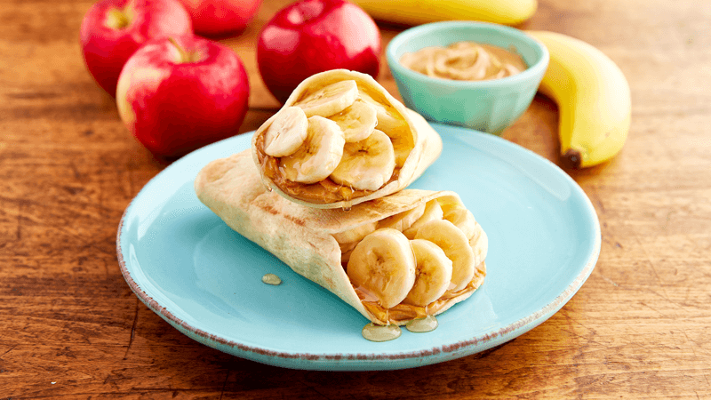 Peanut Butter Banana Tortilla Pockets served in a light blue plate with apple & bananas around on a wooden table